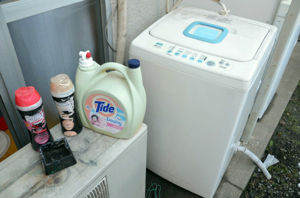 洗濯機置き場が室内になく、雨ざらしの外に置いている。左のボトルは洗濯物の香りづけ。